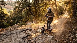 n adult rider navigating a beige NAVEE UT5 Ultra X all-terrain electric scooter over broken pavement and dirt on a scenic, sunny path