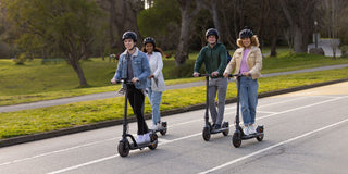 Four riders commuting on lightweight electric scooters along a city park road.